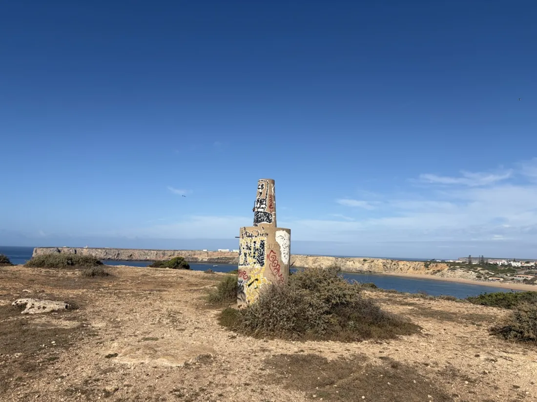 Ein mit Graffiti bedeckter Betonpfeiler auf dem östlichen Kliff, dahinter über der Bucht die flache Landzunge von Sagres